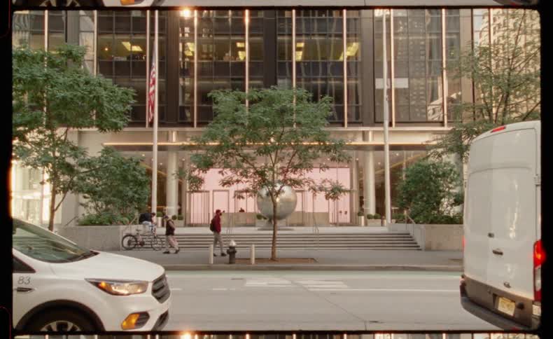 A static, film-style shot captures pedestrians walking along a sidewalk in front of a modern office building in New York City