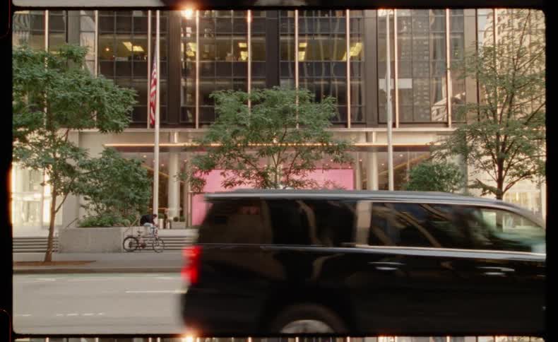 A film-style shot shows pedestrians and traffic passing a modern New York City building with a silver sphere sculpture and pink lit entrance