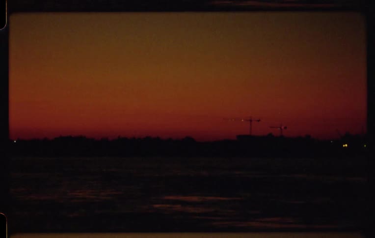 Vintage Super 8 footage of a deep red and orange sunset over a silhouetted industrial shoreline
