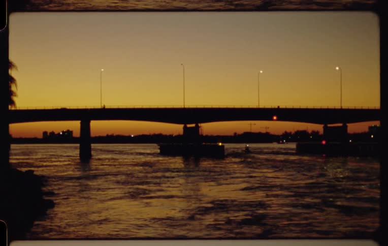 Boats travel under a silhouetted bridge on a river during a golden sunset, captured in a vintage film style