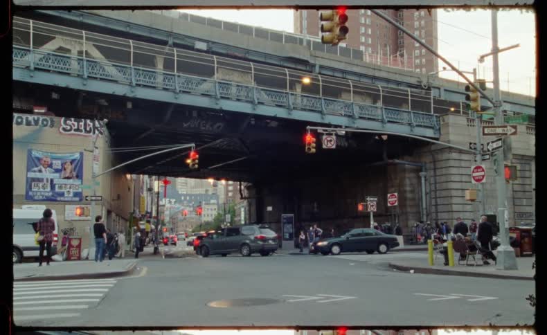 A vintage film-style shot captures people in face masks crossing a busy New York City street under an elevated train