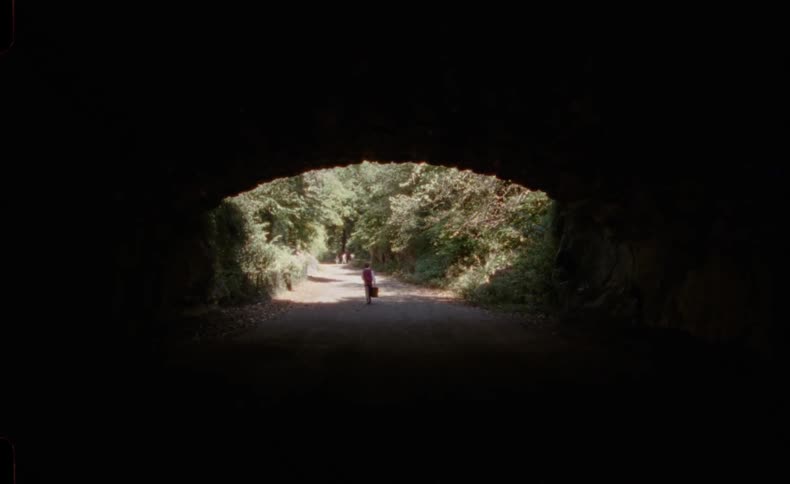 A silhouetted figure walks out of a dark tunnel into the sunlight on a path through the woods