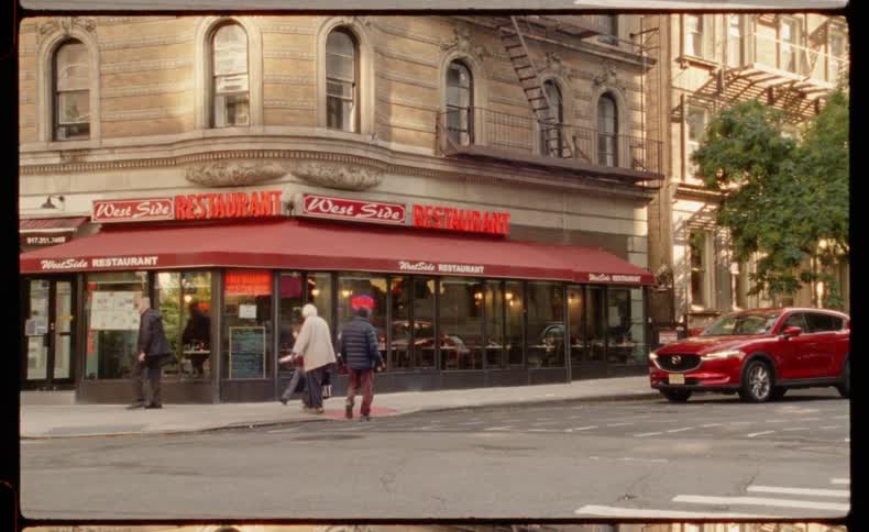 A vintage film-style shot captures daily life on a New York City street corner in front of the West Side Restaurant as people and cars pass by