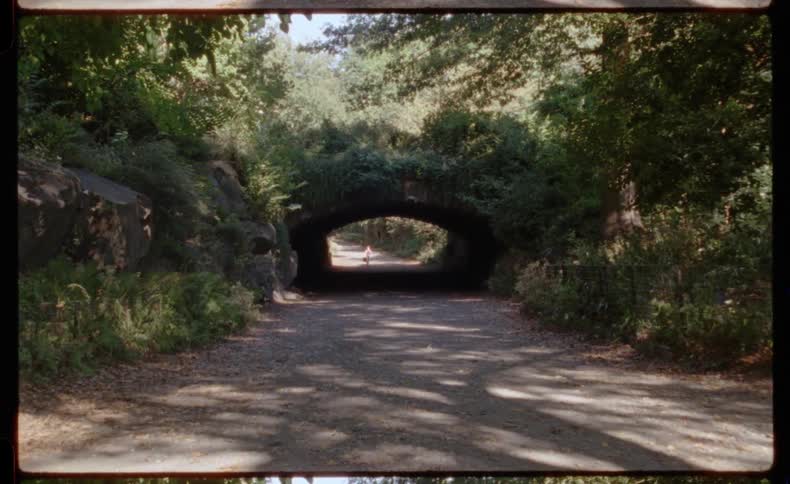 A vintage film shot captures a person walking into a dark, stone tunnel on a sun-dappled park path as a cyclist emerges