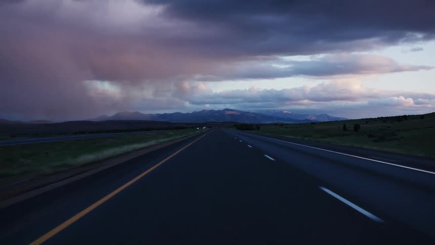 Driving on a vast highway towards distant mountains under a dramatic stormy sky at twilight