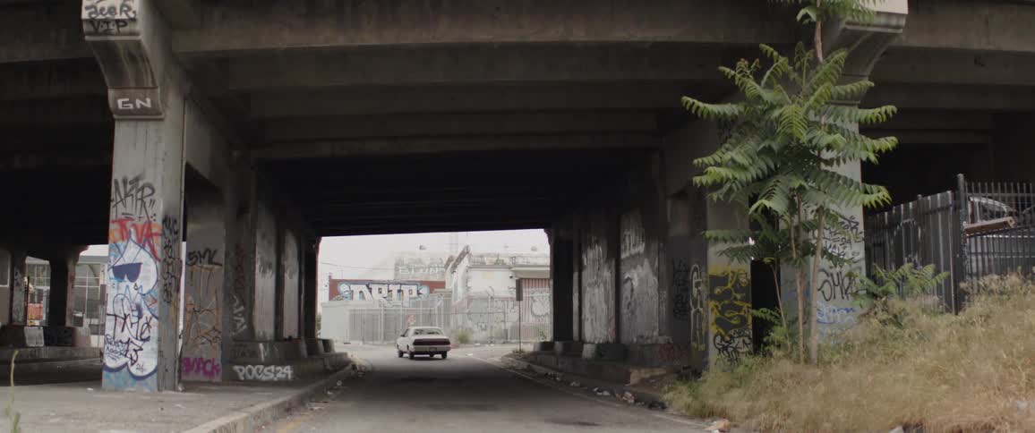 A white classic sedan drives away under a graffiti-covered urban overpass