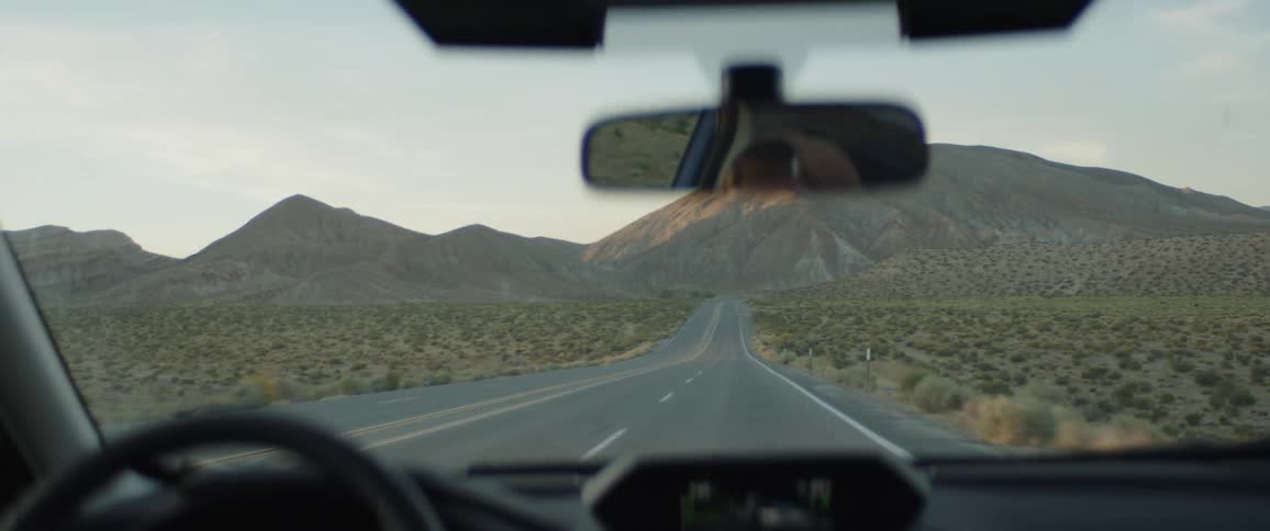 A first-person view from a car driving down a long, empty highway through the desert towards distant mountains