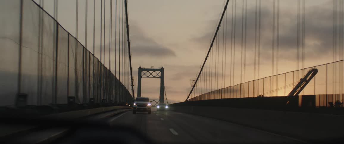 A point-of-view shot from a car driving across a large suspension bridge during a warm sunset