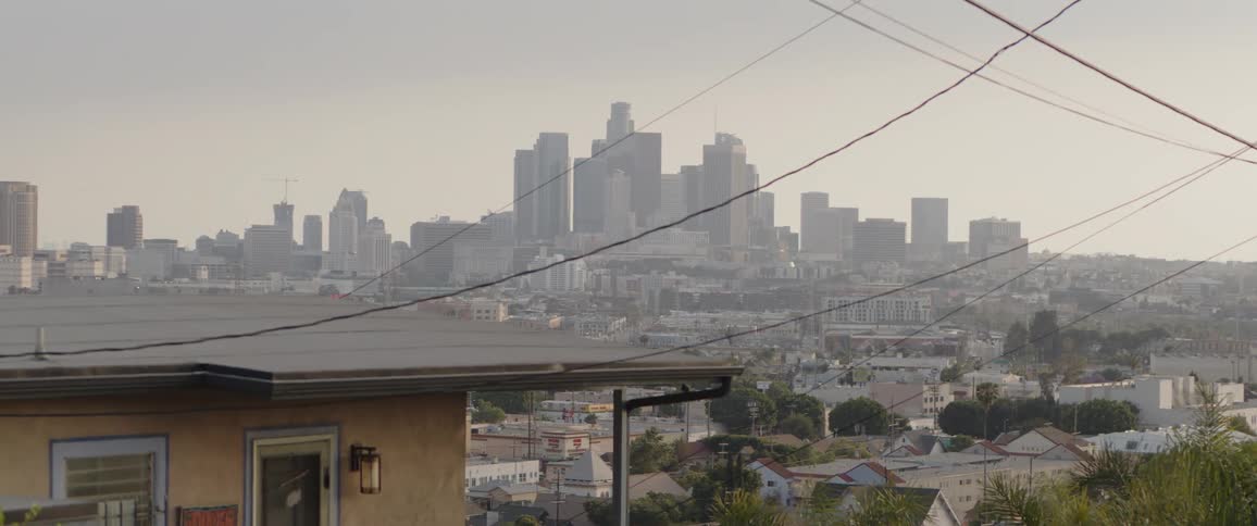 A gritty, hazy view of the downtown Los Angeles skyline slowly zooms out from a suburban vantage point with power lines in the foreground