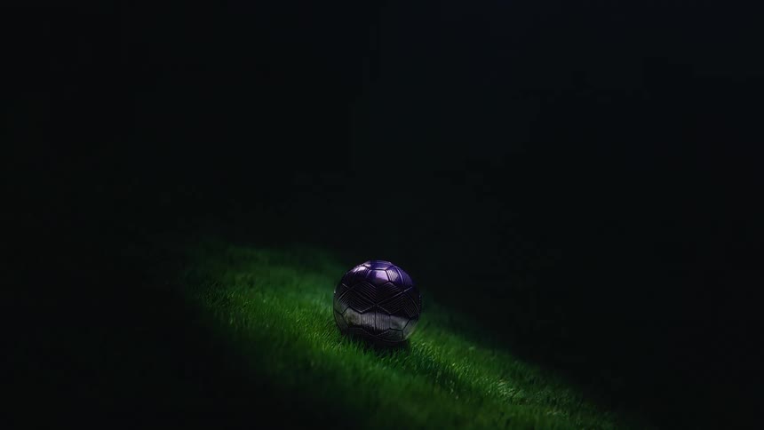 A single metallic soccer ball on a green pitch is dramatically revealed by a moving spotlight