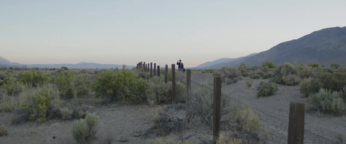 A lone figure sits on a fence post in the vast, empty desert at dusk, looking out at the distant mountains through binoculars