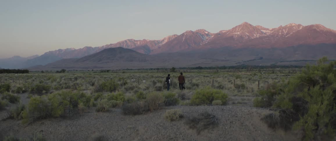 Two people walk through a vast desert valley towards a majestic snow-capped mountain range at sunset