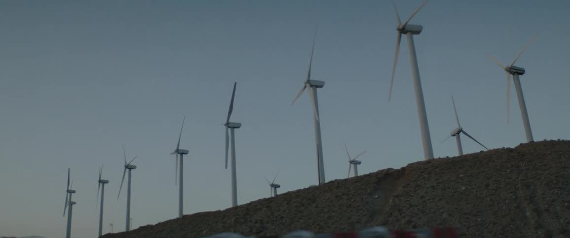 Slow motion view from a car driving past a wind farm with turbines spinning on a desert hill at dusk