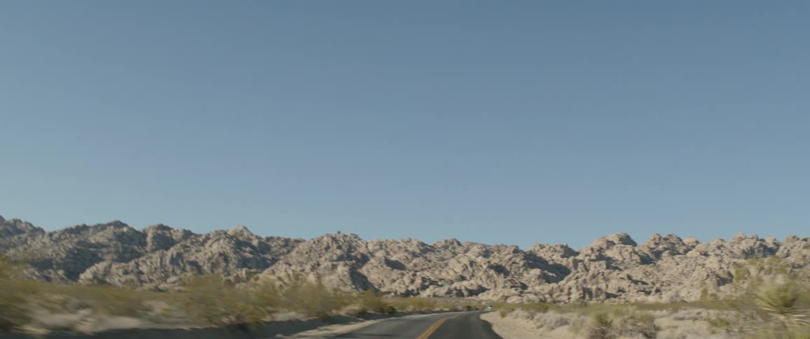 A scenic point-of-view drive through a vast desert landscape with rocky hills under a clear blue sky