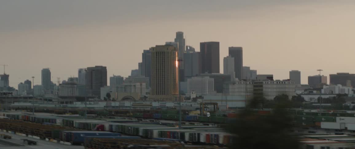 A moving shot reveals the downtown Los Angeles skyline at sunset behind a bustling industrial train yard