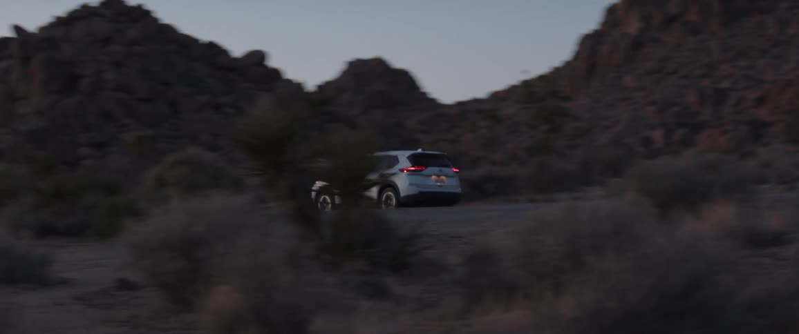 A white SUV drives along a dirt road through a rocky desert landscape with Joshua trees during twilight