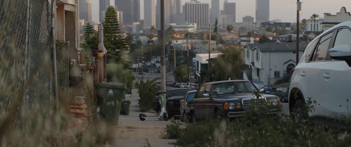 A quiet, overgrown residential street sidewalk with parked cars and an electric scooter has a view of the downtown Los Angeles skyline