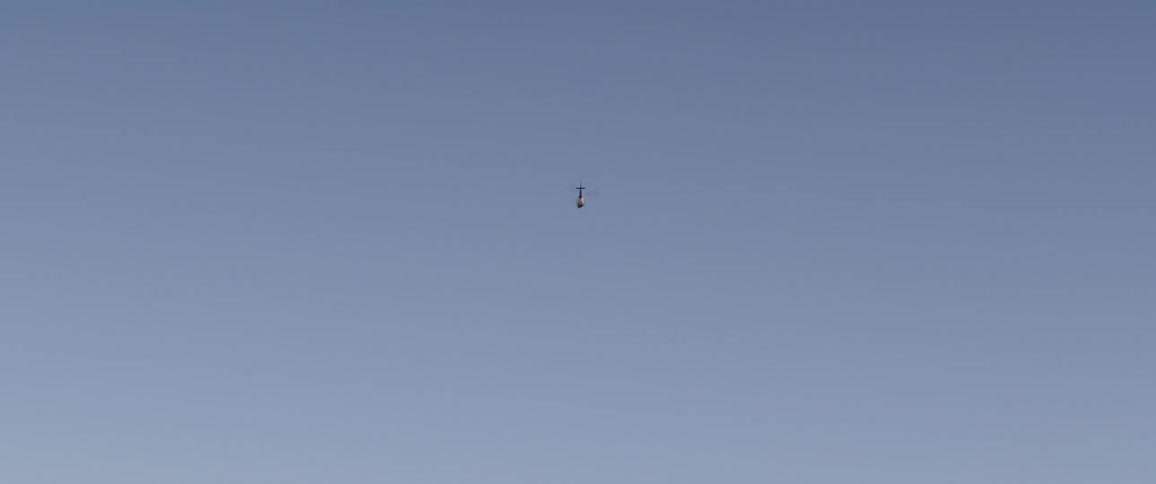 A slow zoom-out shot captures a helicopter flying away and disappearing into the vast, clear blue sky
