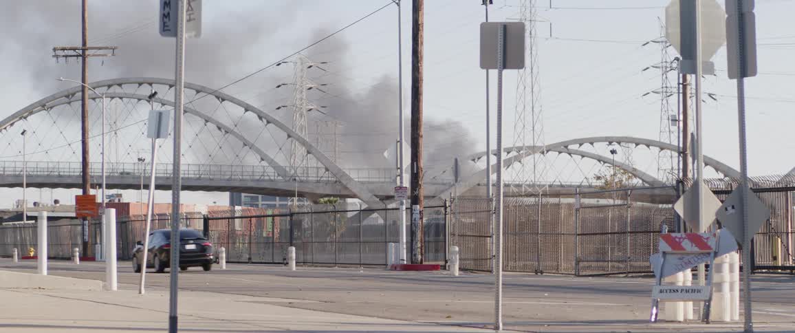 Dark smoke billows from a fire behind the Sixth Street Viaduct in Los Angeles as a car drives by on the street below