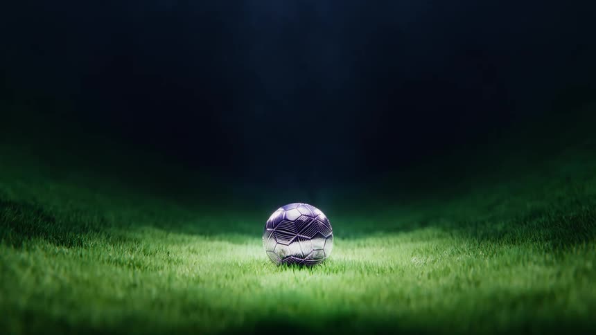 A metallic soccer ball descends in dramatic slow motion, landing in a single spotlight on a dark, grassy field