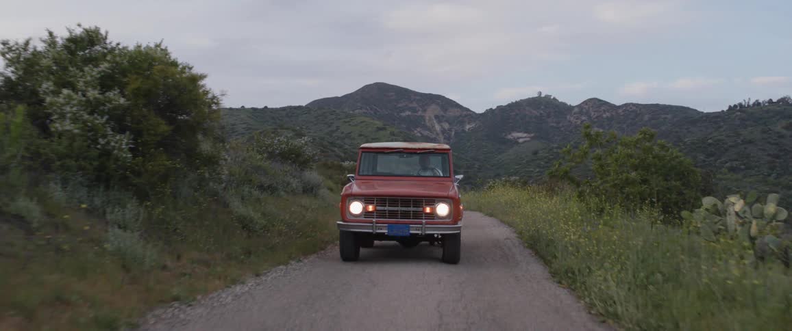 A vintage red SUV with its headlights on drives down a scenic, winding mountain road at dusk