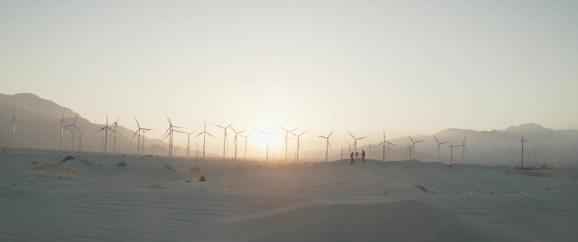 Driving past a sprawling wind farm in a sandy desert landscape as the sun sets on the horizon