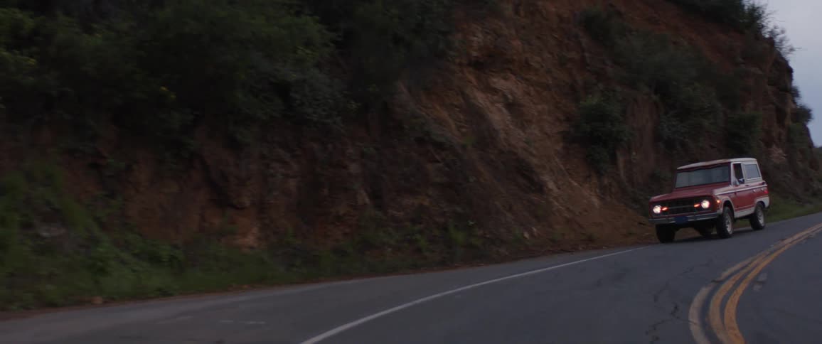A classic red SUV with its headlights on drives in slow motion on a winding mountain road at dusk