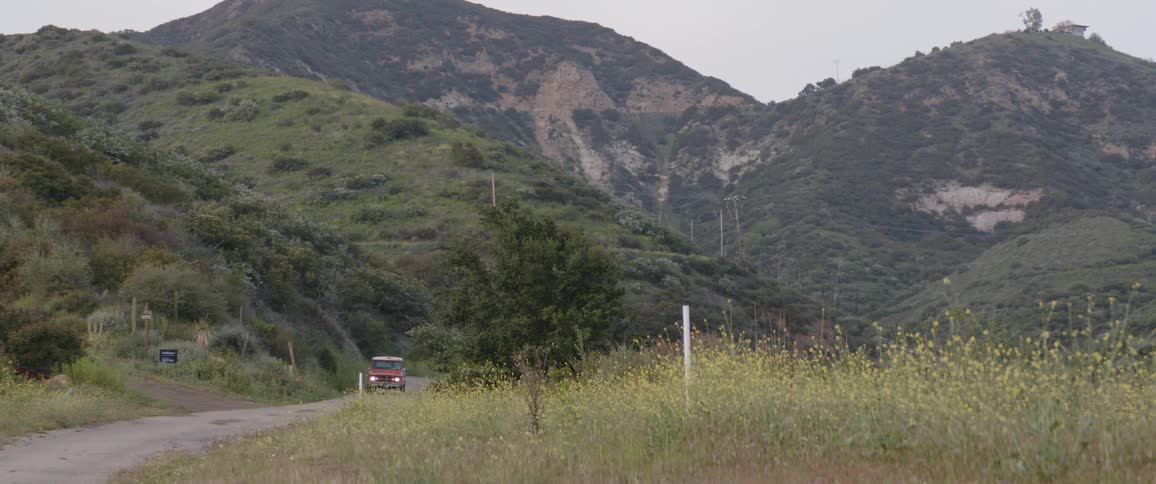 A vintage orange truck drives up a winding road through a lush green valley in the mountains