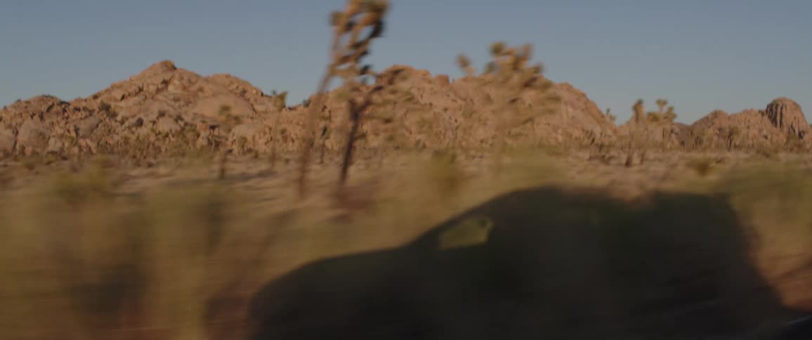 POV from a car driving through the desert landscape of Joshua Tree at sunset, with the vehicle's shadow visible on the ground