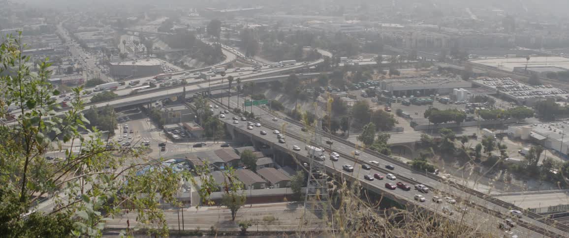 A high-angle view of a busy freeway interchange with heavy traffic flowing through a hazy, smoggy cityscape