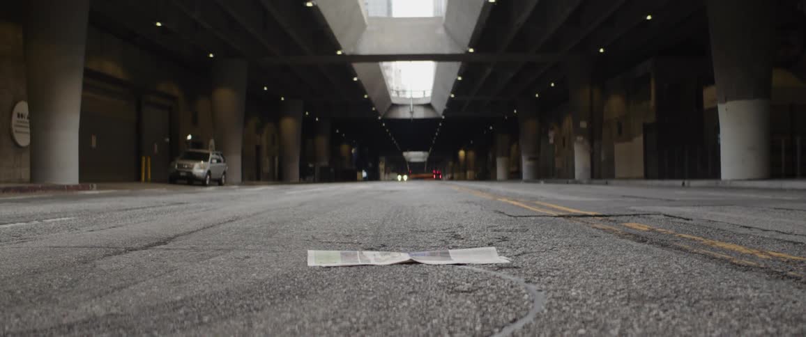 A low-angle shot tilts up from a discarded newspaper on an empty urban street, revealing the concrete architecture of a massive city underpass
