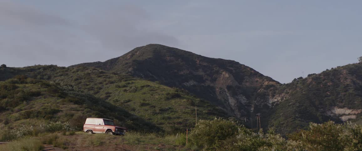 A vintage orange SUV sits parked on a grassy hill against the backdrop of a large, rolling mountain