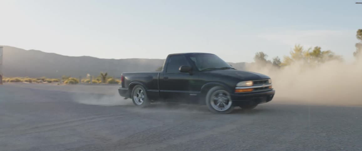 A black pickup truck drifts through a desert lot in slow motion, kicking up a massive cloud of dust against the setting sun