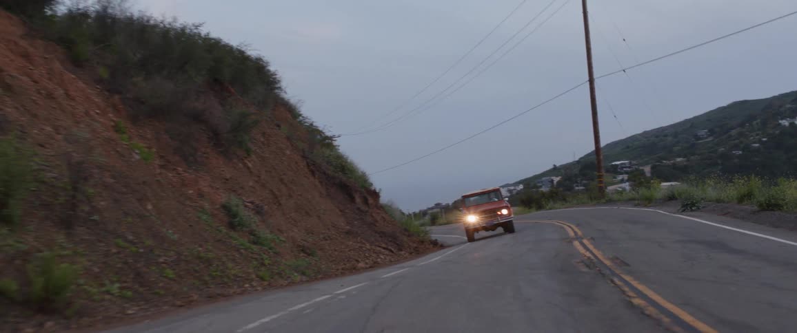 A vintage red truck with its headlights on navigates a winding country road through the hills at dusk