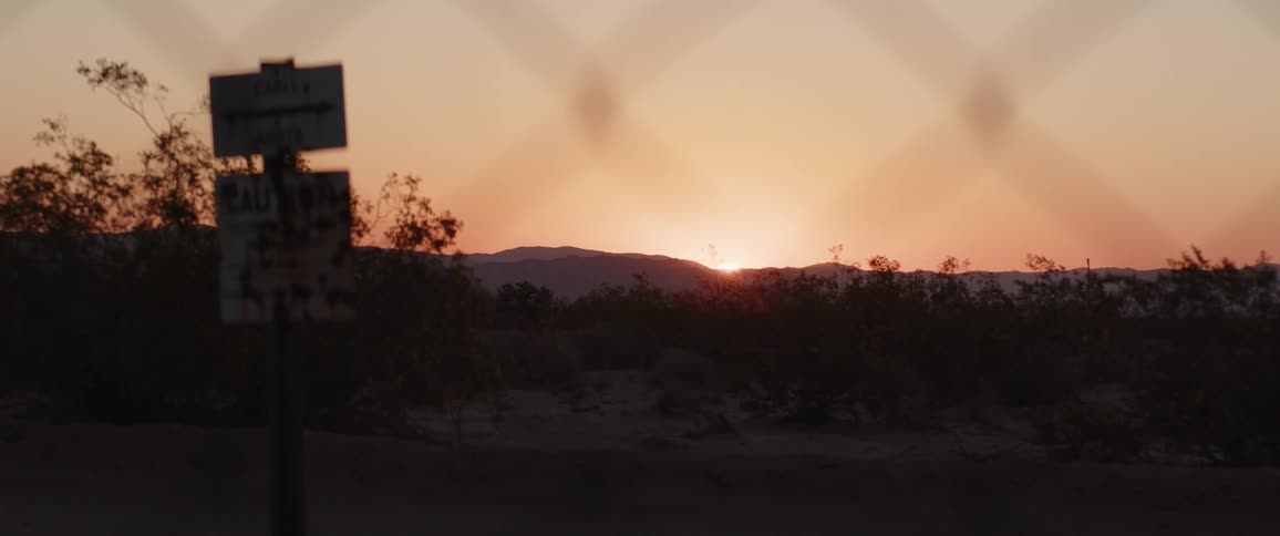 The sun sets over a desert mountain range, viewed through a chain-link fence with a bullet-riddled sign silhouetted in the foreground