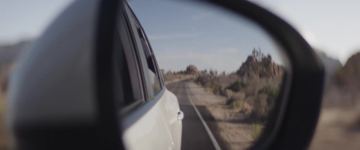 A scenic desert road and rocky landscape are seen in the reflection of a moving car's side mirror