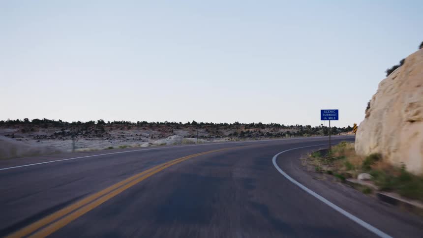 A point-of-view shot driving along a winding desert highway past a rocky bluff at twilight