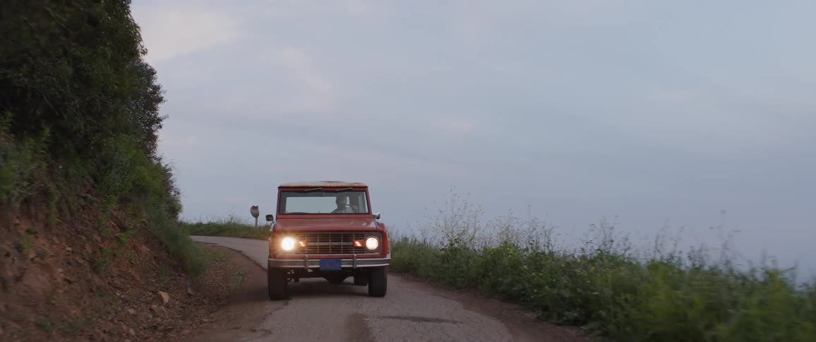 A classic red SUV with its headlights on slowly drives down a winding country road through the hills at dusk