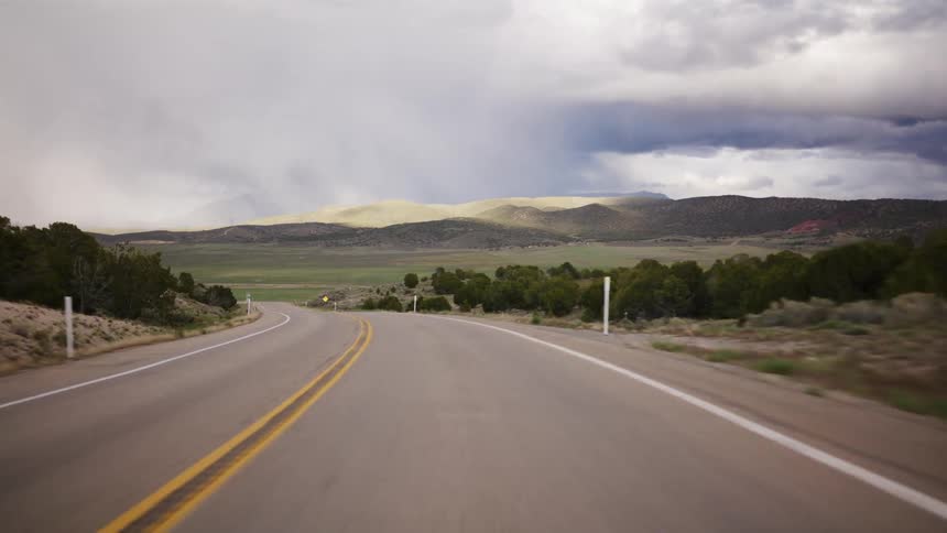 Driving point-of-view on a winding highway through a vast, rural landscape under dramatic, stormy skies