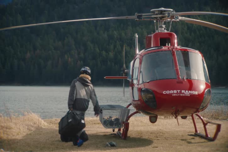 A person in winter gear unloads their bags next to a red heli-skiing helicopter parked on the shore of a mountain lake