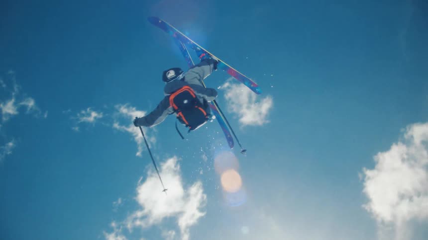 A professional skier performs a spectacular mid-air trick in slow motion against a bright blue sky