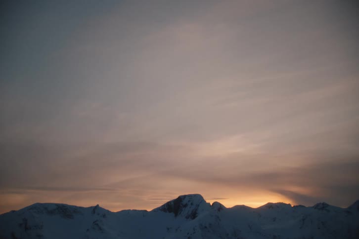 A static shot shows a beautiful, warm sunset behind a vast, snow-covered mountain range