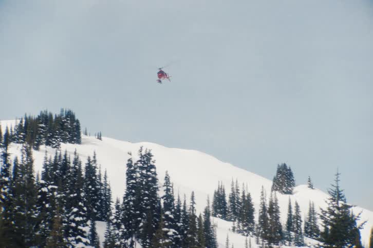 A red helicopter flies low over a snow-covered mountain landscape dotted with evergreen trees
