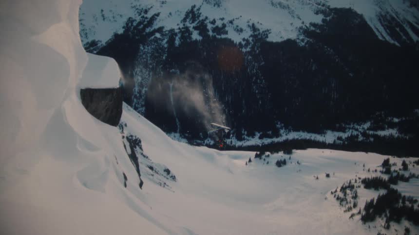 A freestyle skier launches off a steep cliff in slow motion, executing a perfect backflip against a stunning mountain backdrop