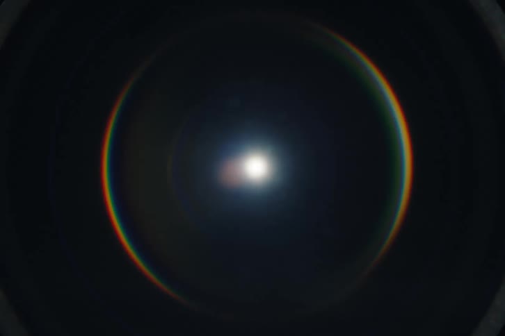 A central point of light creates a slowly expanding circular rainbow lens flare against a black background