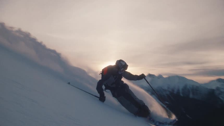 An epic slow-motion shot follows a backcountry skier as he carves a turn through deep powder snow against a stunning mountain sunset