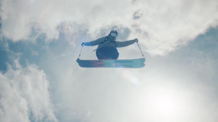 An expert skier soars through the air in slow motion after launching off a snowy jump on a bright sunny day