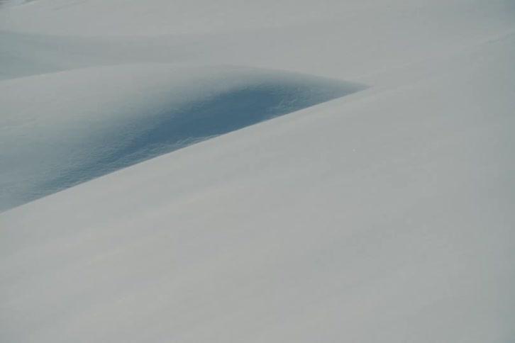 Fine snow particles drift across a minimalist, serene landscape of untouched snow dunes