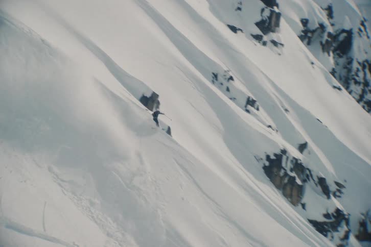 An extreme skier carves through deep powder down a steep and expansive snowy mountain in slow motion