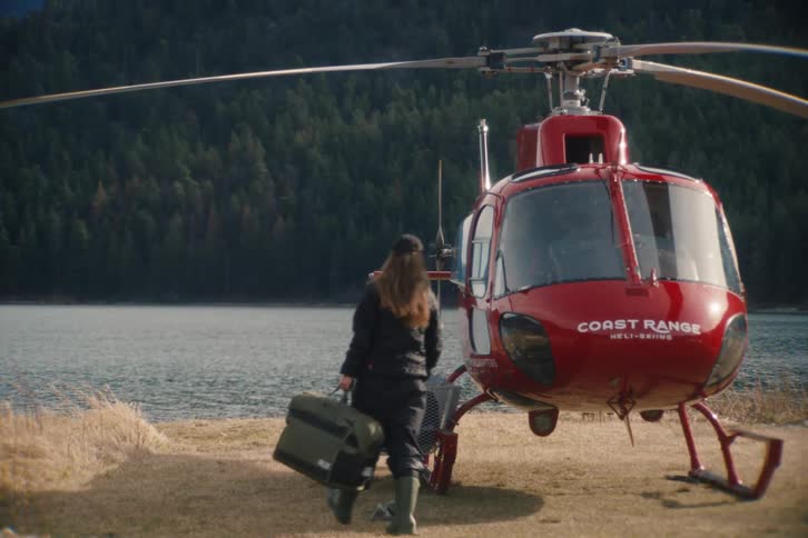 A woman carries her luggage to a red helicopter waiting on the shore of a remote mountain lake